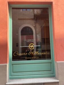 Green-framed glass door with gold lettering that reads “Cravero & Marcialis Scuole di Ballo” in Alba, Italy. The reflection shows a narrow street and arched doorway across the road.
