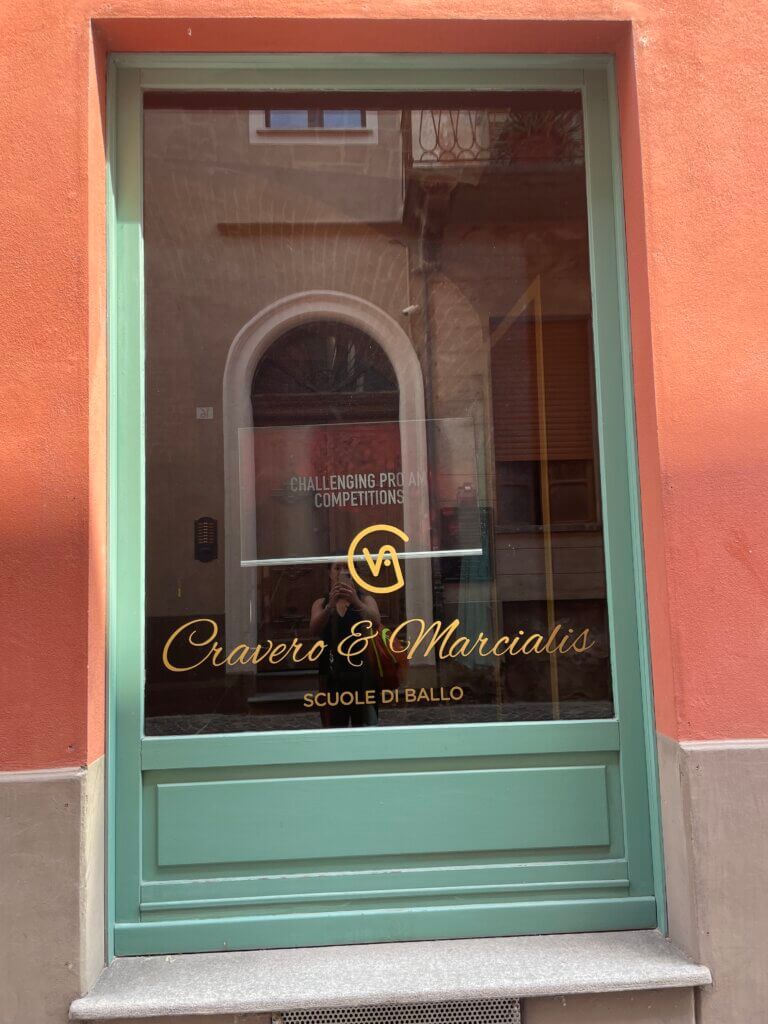 Green-framed glass door with gold lettering that reads “Cravero & Marcialis Scuole di Ballo” in Alba, Italy. The reflection shows a narrow street and arched doorway across the road.