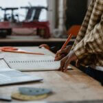 An artisan sketches designs at a workbench in a woodworking shop filled with tools.