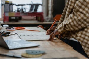 An artisan sketches designs at a workbench in a woodworking shop filled with tools.