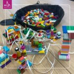 Colourful LEGO® bricks and small constructed towers connected by white strings are spread across a wooden table, with a fabric bowl of loose bricks in the background, illustrating a collaborative LEGO® Serious Play workshop setup.